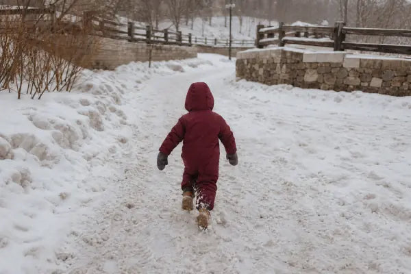 A child in a burgundy snowsuit walks along a snowy outdoor path, seen from behind.
