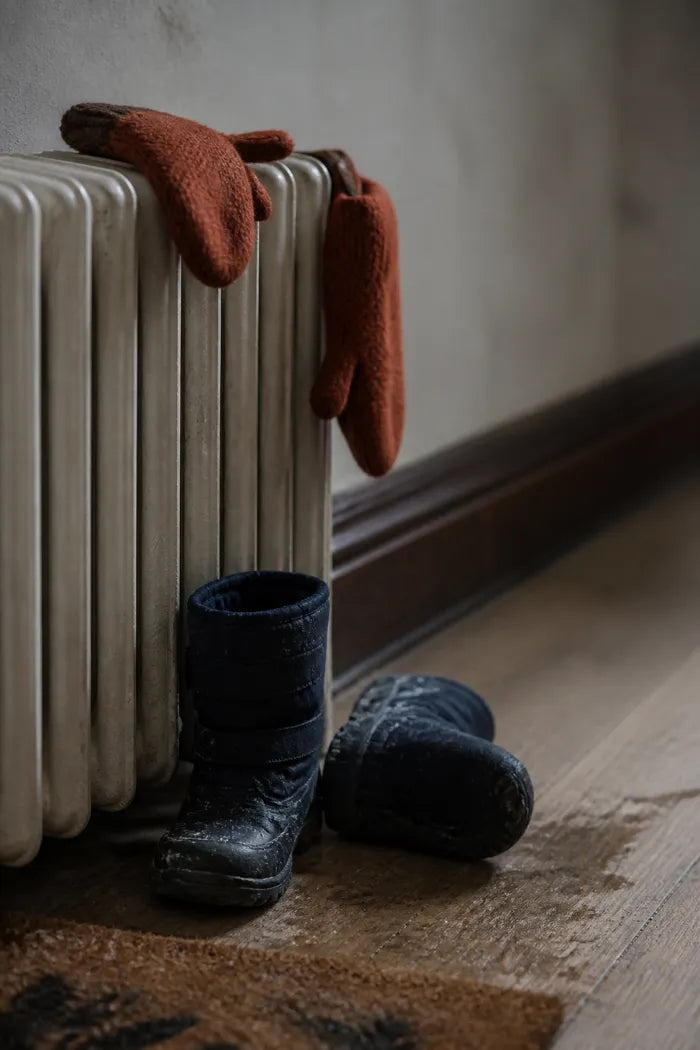 A child's winter boots kicked off at the foot of a radiator, with knitted mittens laid on top of it to dry.
